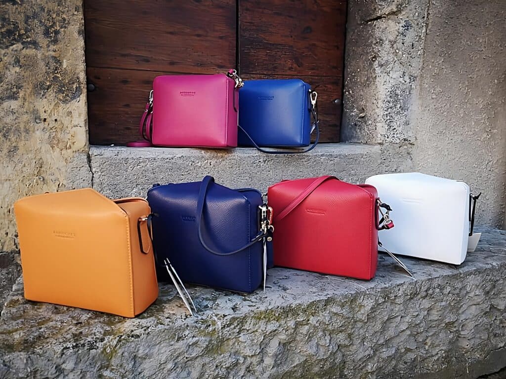 Collection sacs à main cuir femme: couleurs vives et design Six sacs à main en cuir multicolores (fuchsia, bleu, orange LORENZI, rouge, blanc) sur des marches en pierre devant une porte en bois.
