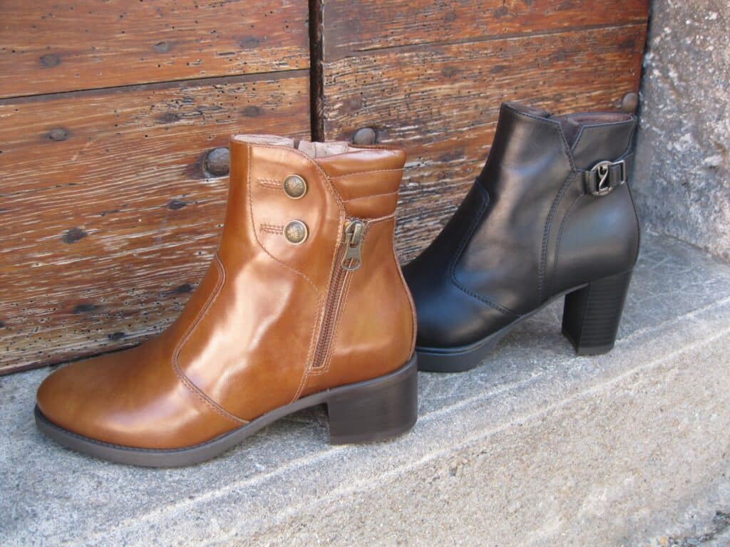 Deux bottines en cuir sur une marche en béton. Une marron clair brillante avec zip YKK et boutons, une noire mate avec boucle. Fond de porte en bois rustique.