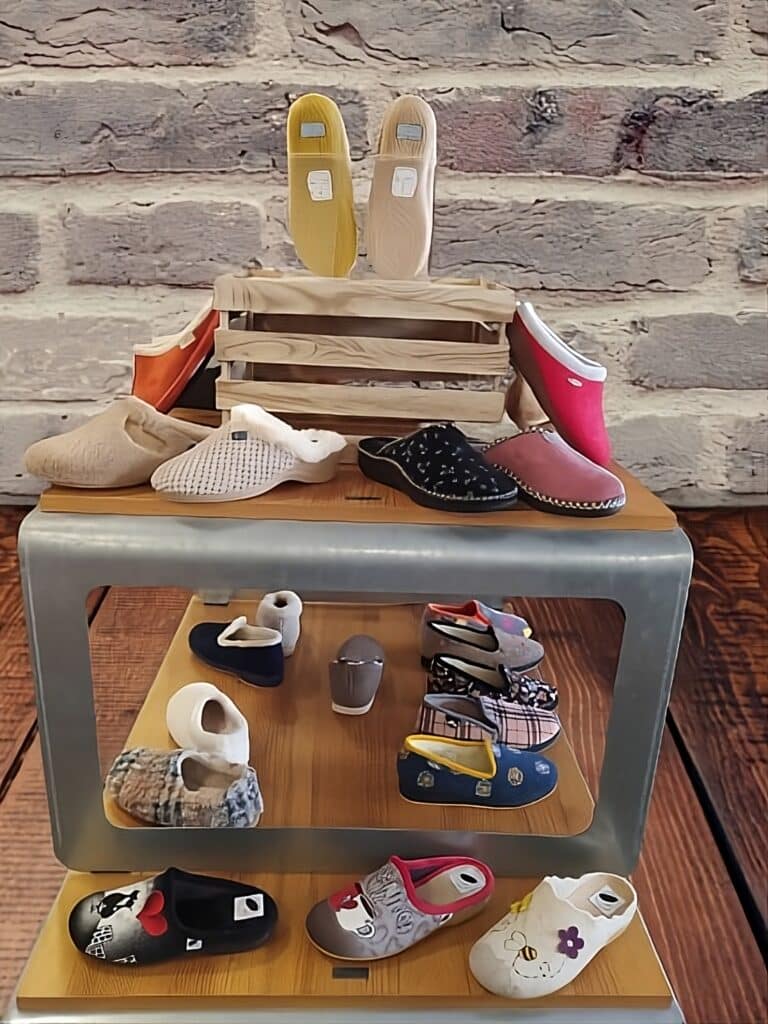 Chaussons & Pantoufles Confortables – Idéal pour la Maison Présentation de nombreux chaussons et pantoufles colorés sur un support, devant un mur de briques et un sol en bois. Idéal confort intérieur.