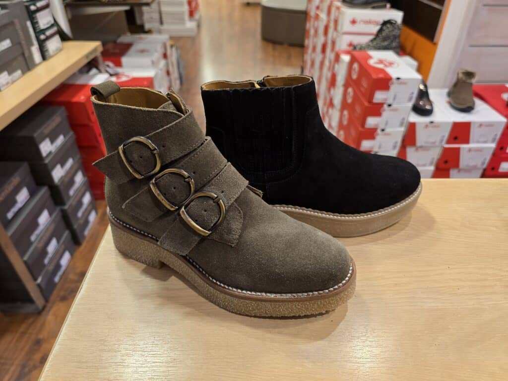 Bottines en Daim Kaki et Noires à Boucles Deux bottines en daim, une kaki à trois boucles et une noire style Chelsea, posées sur un comptoir en bois, avec des boîtes à chaussures Ri**ker en arrière-plan.