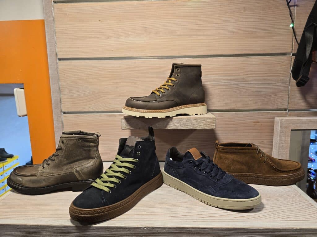Quatre paires de chaussures pour hommes exposées sur des étagères en bois clair : une botte marron foncé, une bottine kaki, une sneaker bleu marine à lacets verts et une autre sneaker bleu marine, avec une étiquette 'keb' visible.